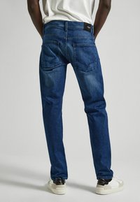 Pantalones vaqueros de mezclilla azul con corte recto, que cuentan con bolsillos traseros y una etiqueta oscura en la cinturilla. Llevados con zapatillas blancas.