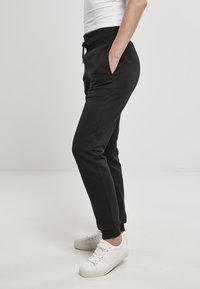 Schwarze Jogginghosen mit elastischem Bund, Seitentaschen und schmal zulaufenden Beinabschlüssen. Kombiniert mit weißen, niedrigen Sneakern. Glatte Stofftextur.
