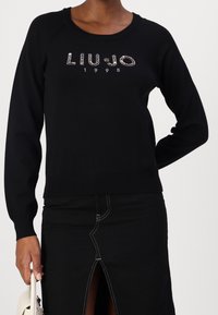 Personne portant un pull noir à manches longues avec l'inscription en strass "LIU JO 1995" et une jupe noire fendue à l'avant, tenant un sac à main blanc.