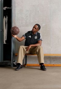 Muže sedí na dřevěné lavici a točí basketbalovou míč na prstu vedle otevřené skříňky s sportovním oblečením a botami proti betonové zdi.