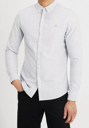 Hombre vistiendo una camisa blanca de manga larga, entallada, con finas rayas verticales grises y pantalones negros, de pie con una mano en el bolsillo.