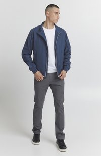 Marineblå letvægtsjakke med lynlåsfront, ståkrave og elastiske manchetter, parret med grå bukser og sorte sneakers.