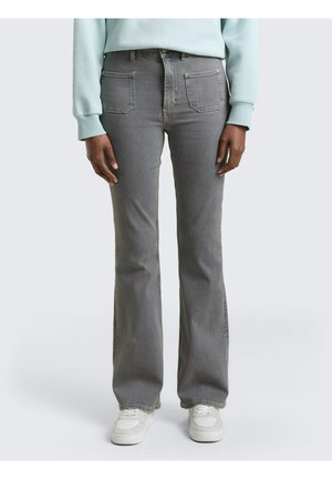 Persoon die grijze recht toelopende spijkerbroek, lichtblauwe trui en witte sneakers draagt, staand tegen een effen lichte achtergrond.