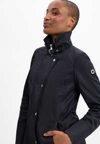 Schwarze wasserdichte Jacke mit hohem Kragen, silberner Hardware und Druckknöpfen. Verfügt über seitliche Taschen und eine taillierte Passform. Keine sichtbaren Muster.