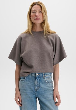 Donna bionda che indossa una maglietta grigia larga a maniche corte e jeans azzurri a vita alta, in piedi su uno sfondo bianco uniforme.