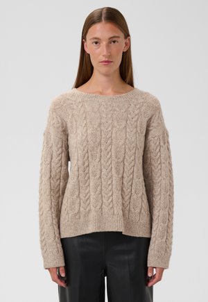 Femme aux cheveux bruns et raides portant un pull beige à torsades et un pantalon noir, debout devant un fond gris clair.