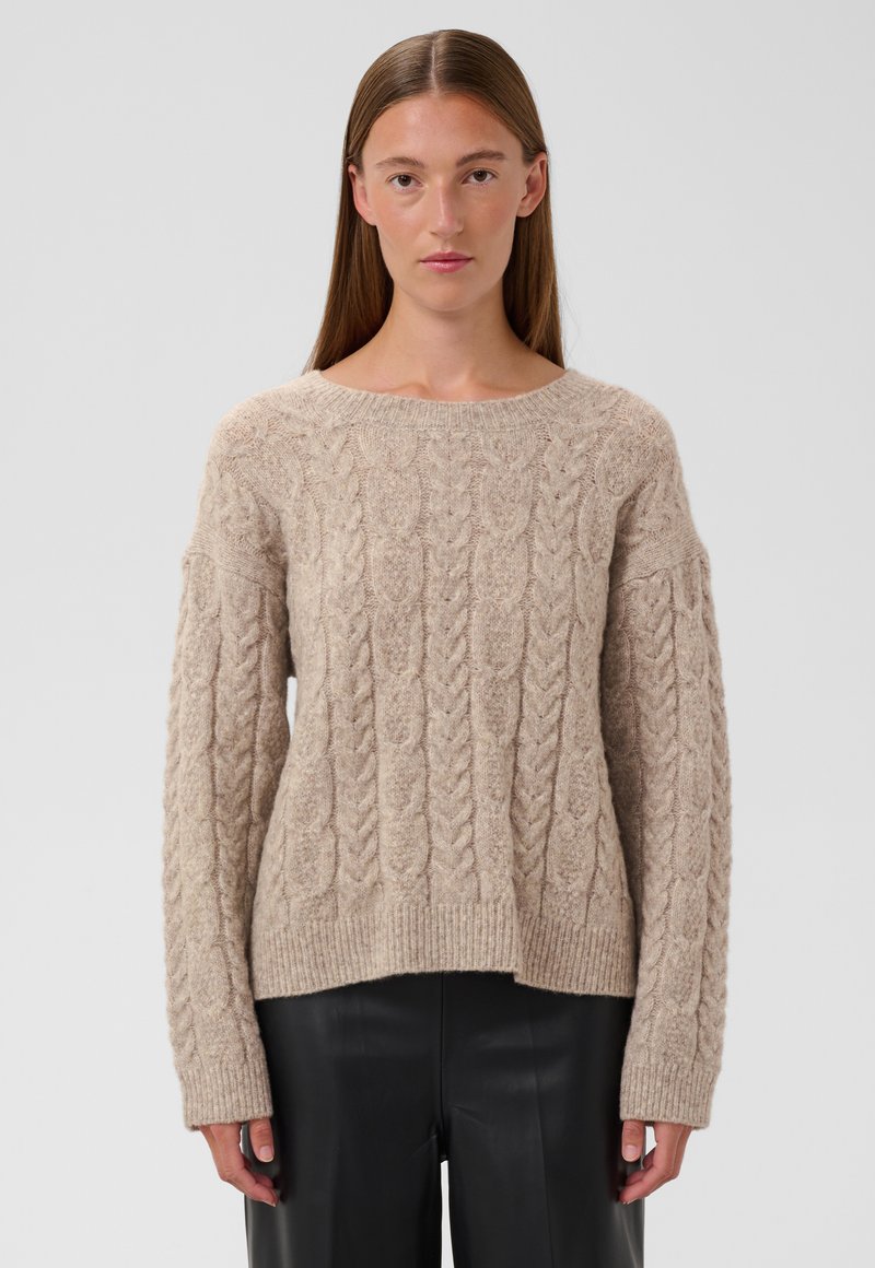 Femme aux cheveux bruns et raides portant un pull beige à torsades et un pantalon noir, debout devant un fond gris clair.