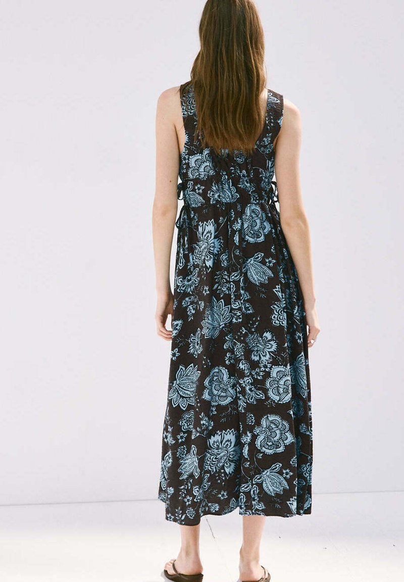 Femme aux longs cheveux bruns portant une robe noire sans manches avec des motifs floraux bleus et des sandales, debout de dos sur un fond uni.