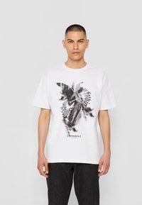 Hvid bomulds T-shirt med et sort-hvidt grafisk print af en engel og kyrillisk tekst. Korte ærmer og en afslappet pasform.