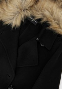 Melnā vilnas mēteli ar mākslīgā kažoka apkakli, pielāgotiem klapjiem un pogu aizdari. Gluda tekstūra ar minimālu redzamu šuvju detaļu.