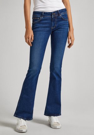 Person iført blå flare jeans med hvide sneakers og en hvid ærmeløs top, stående mod en ensfarvet lys baggrund.