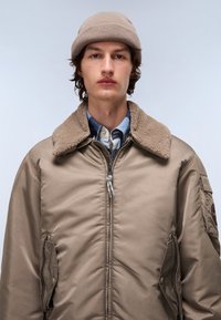 Beżowa ocieplana kurtka z kołnierzem z owczej wełny, zamek błyskawiczny, boczne kieszenie i naszywka na rękawie. Nosi ją na niebieskiej flanelowej koszuli i dzianej czapce.