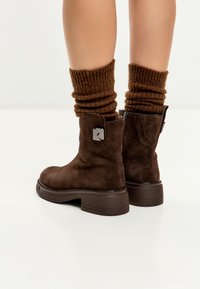 Stivali alla caviglia in suede marrone con zip laterale e dettaglio metallico, indossati con calze marroni a coste spesse su gambe nude su uno sfondo bianco.