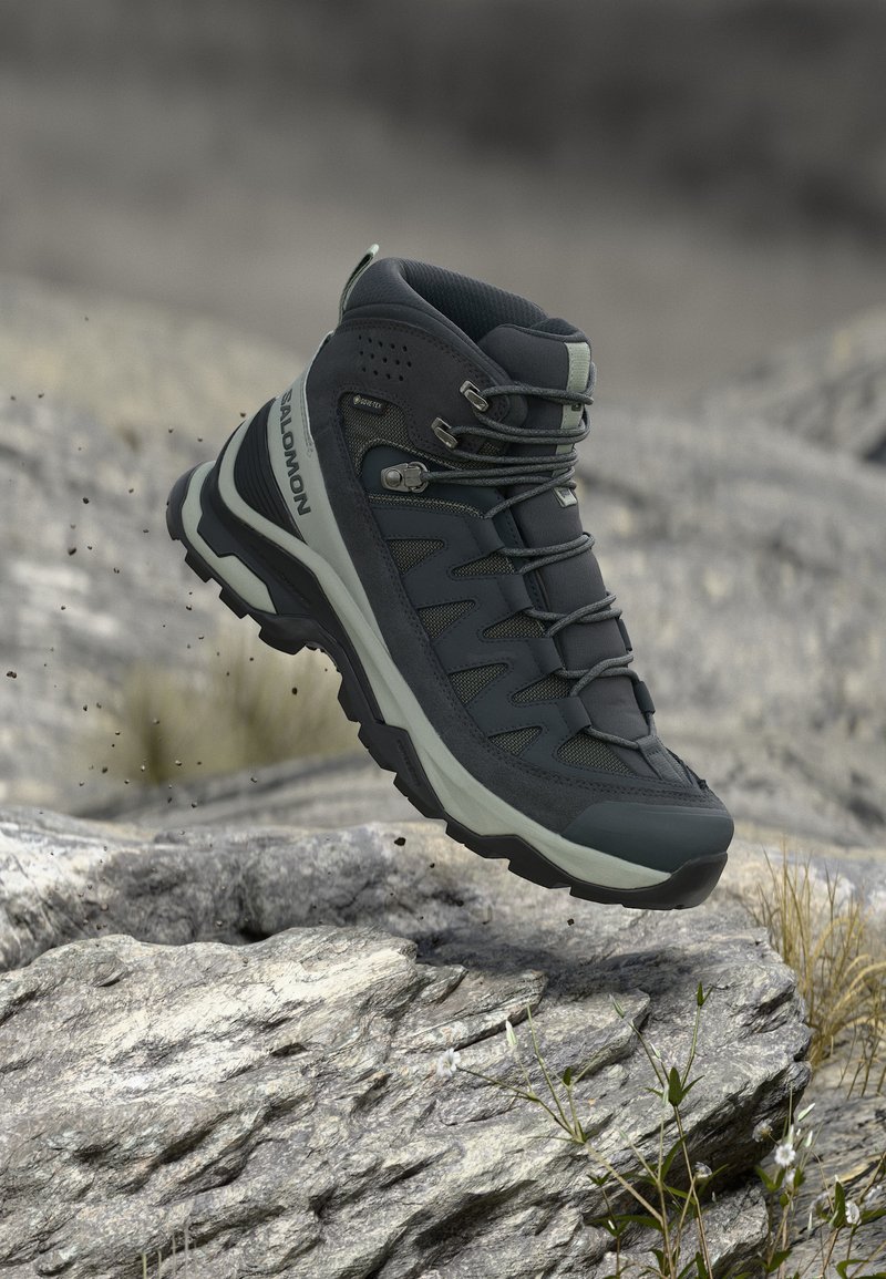 Μαύρο και γκρι μπότες πεζοπορίας με κορδόνια που αιωρούνται πάνω από βραχώδη έδαφος με μικρά φυτά και χαλίκια σε φυσικό υπαίθριο περιβάλλον.