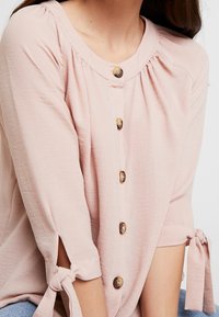 Blusa rosa chiaro con scollatura rotonda e quattro bottoni sul davanti. Presenta maniche corte con fiocchi e delicate arricciature sulle spalle.