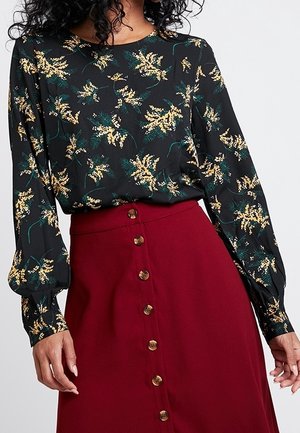 Femme portant un blouse floral à manches longues noire rentré dans une jupe boutonnière bordeaux taille haute.