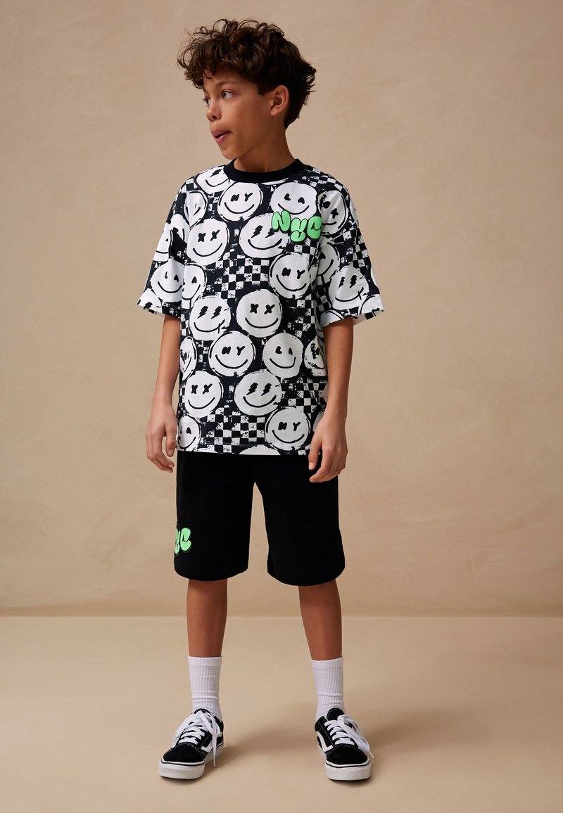 T-shirt graphique noir et blanc à manches courtes avec des visages souriants et un motif damier, accompagné d'un short noir avec un détail de logo vert.
