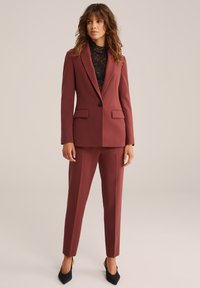 Burgundska po meri krojena blazer jakna z usklajenimi hlačami, ki vključuje ostre reverje, dvoje sprednjih žepov in en sam črn gumb. Pod njo je črna čipkasta bluza.