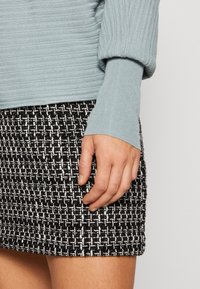 Gros plan sur une main reposant à la taille, vêtue d'un pull bleu clair côtelé et d'une jupe en tweed à motifs noir et blanc.