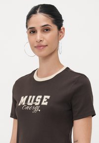 Rudas medvilninis marškinėliai su grietinėlės spalvos raukšlėtu apykaklės kraštu. Yra atspaustas "MUSE Energy" logotipas baltos ir grietinėlės spalvų su stilizuotu dizainu.