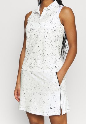 Femme portant un haut blanc sans manches à col et une jupe assortie avec de petits pois noirs et des logos Nike, main dans la poche de la jupe.