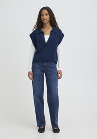 Pull sans manches tricoté bleu marine avec col en V et col, associé à un jean droit bleu et des chaussures noires sans lacets.
