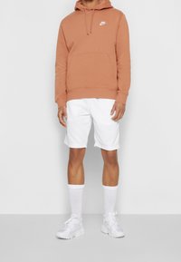 Mikina Nike v barvě zaprášené oranžové s klokánkovou kapsou, doplněná bílými šortkami. Doplňky zahrnují bílé ponožky a světlé tenisky.