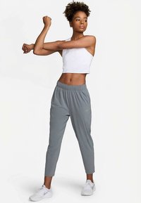 Szürke sportnadrág elasztikus derékpánttal és testhez simuló kialakítással, fehér, rövidített ujjú toppal párosítva. A modell a karjait nyújtja.