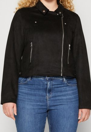 Femme portant une veste de motard noire zippée et un jean bleu, debout devant un fond clair uni.