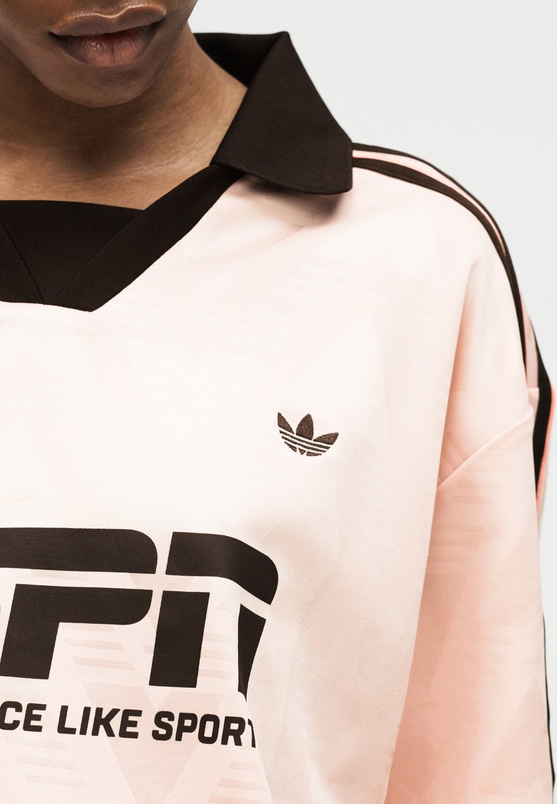 Personne portant un maillot de sport rose pâle avec un col noir, des bandes noires sur les épaules et un texte noir en gras partiellement lisible "SPORT" sur le devant.