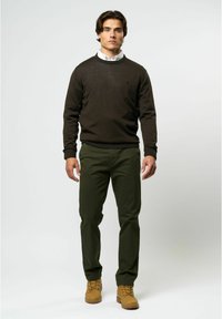 Temno rjava pulover z rebrastimi manšetami, v kombinaciji z zelenimi hlačami in svetlo rjavimi škornji. Obleka vključuje belo srajco s ovratnikom spodaj.