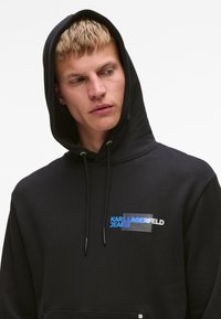 Sudadera negra con un bolsillo frontal, con una impresión de logo en azul y blanco. Tejido suave, cordones ajustables y un corte relajado.