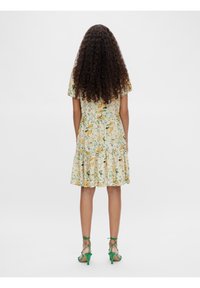 Robe fleurie à manches courtes, motifs jaunes et verts, jupe évasée, tissu texturé. Sandales vertes à talons hauts et à lanières.