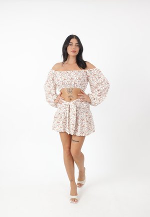 Frau steht und trägt ein weißes geblümtes Off-Shoulder Crop Top und passenden Rock, posiert mit Händen in den Hüften vor weißem Hintergrund.