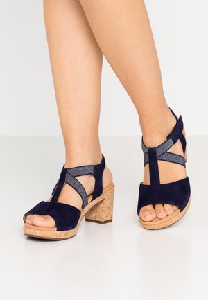 Kvinde iført marineblå ruskind open-toe sandaler med korkblokke og elastiske krydsremme, stående på en hvid overflade.