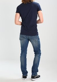 Námořnicky modré tričko s krátkým rukávem, spojené se světle modrými úzkými džínami. Obuv tvoří černé tenisky s bílými podrážkami. Pohled zezadu.