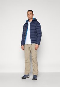 Mornarska pufasta jakna s tirkiznom podstavom i zatvaračem, nosi se preko bijele košulje, u kombinaciji s bež cargo hlačama i sivim planinarskim cipelama.