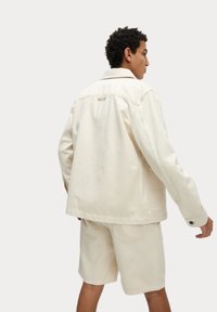 Beige denim jacket with a relaxed fit, buttoned cuffs, and back yoke detail, paired with matching shorts in a similar light color.