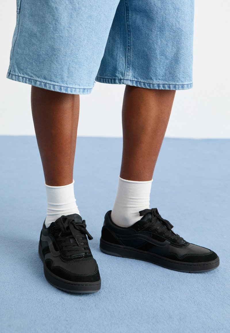 Zapatillas negras con materiales de ante y cuero, que presentan un diseño de cordones, costuras en contraste y una suela de goma texturizada.