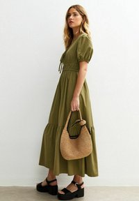 Vestido maxi de algodón color verde oliva con mangas abullonadas y cintura fruncida, combinado con un bolso de mimbre color tostado y sandalias de plataforma negras.