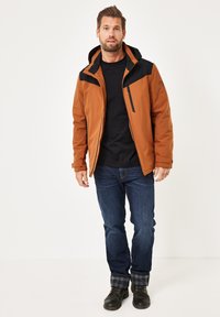 Orangene und schwarze isolierte Jacke mit einer Reißverschlusstasche, getragen über einem schwarzen Hemd. Blaue Jeans und dunkle Stiefel vervollständigen das Outfit.