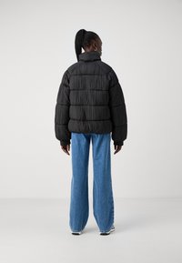 Jachetă puffer neagră cu model matlasat vertical, guler înalt și mâneci lungi. Asortată cu blugi din denim albastri cu pantaloni largi.