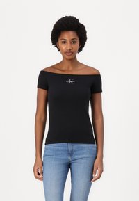 Juodas off-the-shoulder tvirtas mar�škinėliai, pagaminti iš medvilnės, su mažuoju baltu Calvin Klein logotipu ant krūtinės, derinami su šviesiai mėlynomis džinsais.