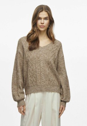 Pull en tricot beige avec un col en V, motifs en cable texturés, et manches ballon. Poignets et ourlet côtelés, associé à un pantalon de couleur claire.
