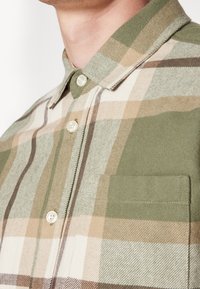 Gros plan d'une personne portant une chemise à carreaux beige, verte et marron avec boutons, une poche avant et un col.