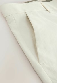 Pantalones de algodón beige con una textura suave, que presentan un pliegue limpio en la cinturilla y detalles de costura sutiles.