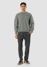 Sweatshirt gris avec col rond et poignets ribelés, associé à un pantalon plissé gris foncé et à des baskets brunes avec des rayures blanches.