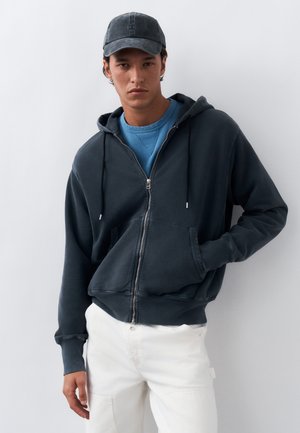 Νέος άνδρας που φοράει σκούρα ζακέτα με φερμουάρ, μπλε φούτερ, μαύρο καπέλο και λευκό παντελόνι, στέκεται μπροστά σε απλό φωτεινό φόντο.