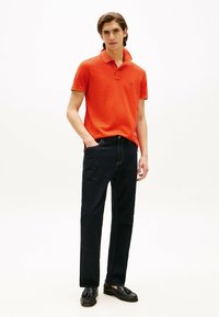 Pomarańczowa koszulka polo z krótkim rękawem, zestawiona z ciemnoniebieskimi dżinsami. Model stoi z jedną ręką w kieszeni, nosząc czarne loafersy.