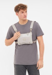 Borsa a petto grigia realizzata in materiale durevole con spallacci regolabili. Presenta una tasca anteriore con zip e un patch con logo. Indossata sopra una maglietta grigia.